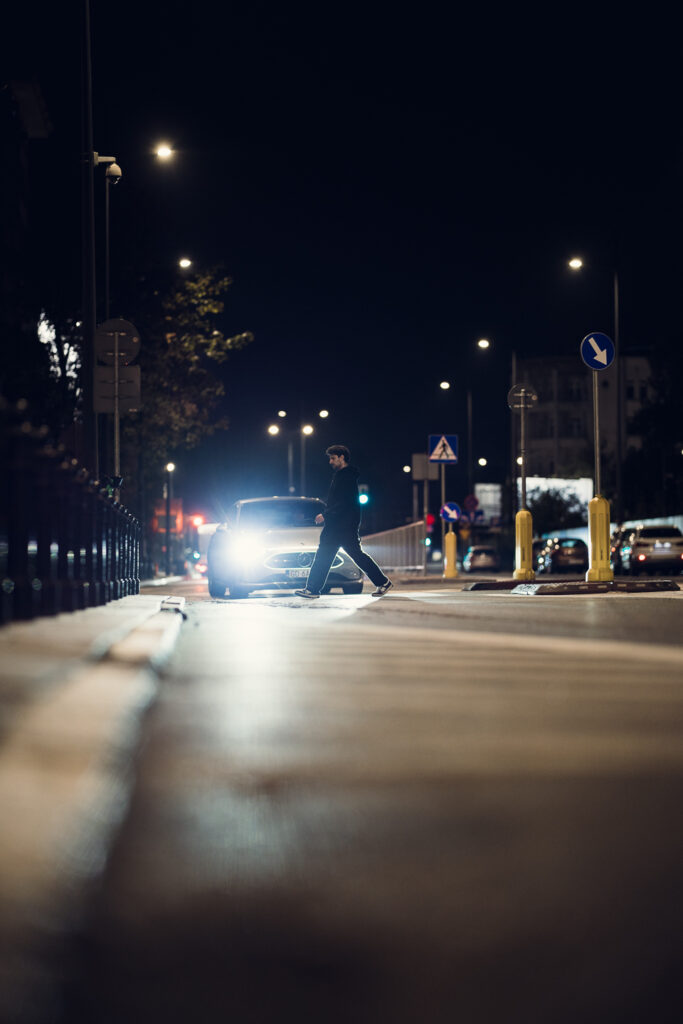 Man crossing a Warsaw street at night, backlit by car headlights, shot low on the Sigma 85mm F1.2 at F1.2 with blurred asphalt in front.