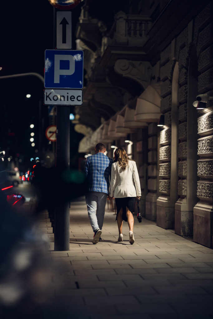 Couple walking hand in hand past an ornate Warsaw facade at night, Sigma 85mm F1.2 isolating them from blurred city lights.