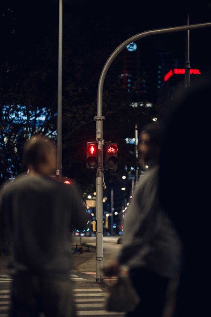 Red pedestrian and bike signals on a Warsaw crossing at night, framed between two out-of-focus pedestrians in the foreground
