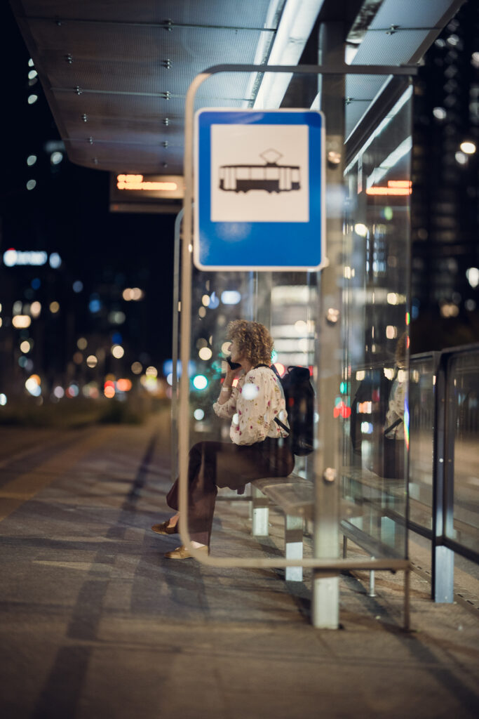 Woman on the phone at a Warsaw tram stop at night, shot through the shelter glass on the Sigma 85mm F1.2 with city bokeh behind