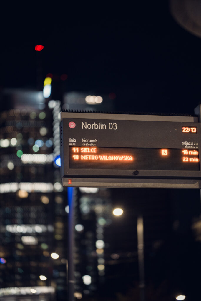 Tram departure board at the Norblin stop in Warsaw at night, sharp against blurred office tower lights