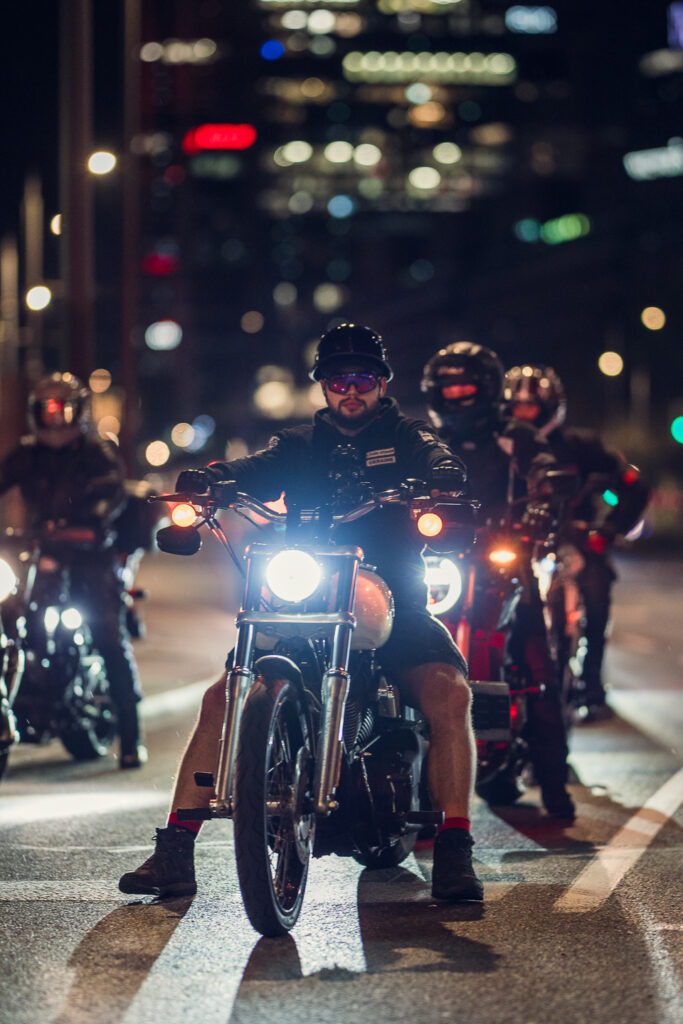 Group of motorcyclists stopped on a Warsaw street at night, the lead rider sharp on the Sigma 85mm F1.2 and the riders behind falling into blur