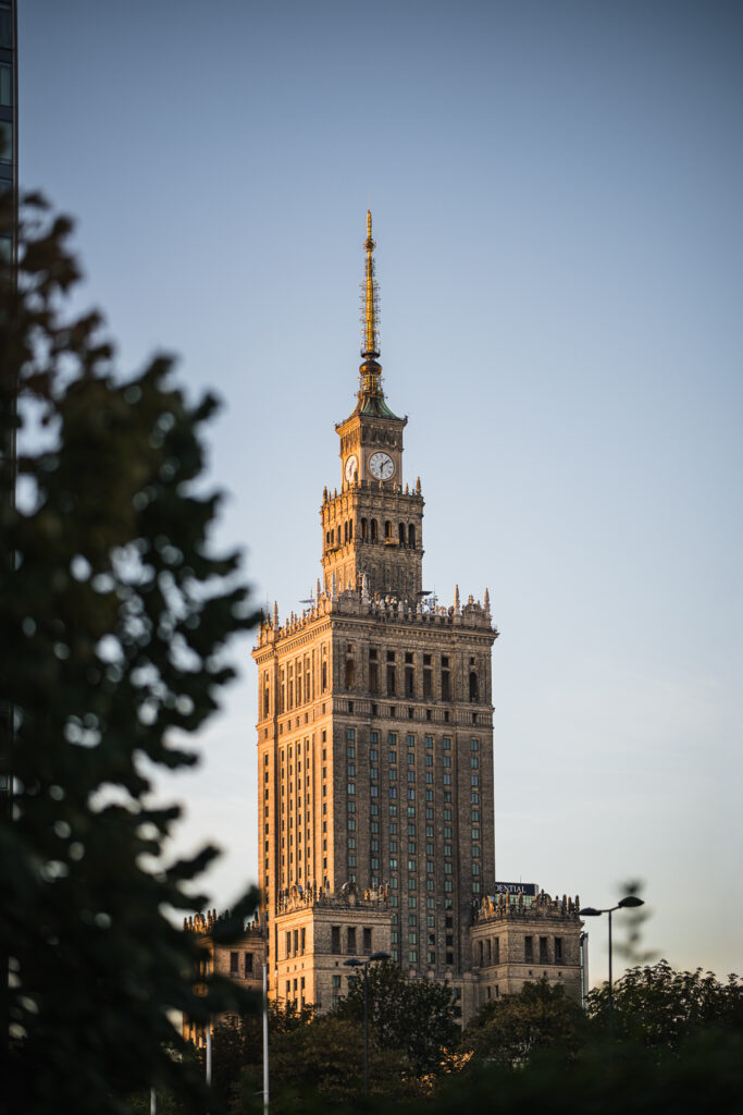 Palace of Culture and Science in Warsaw lit by golden-hour sun, framed by out-of-focus tree leaves in the foreground