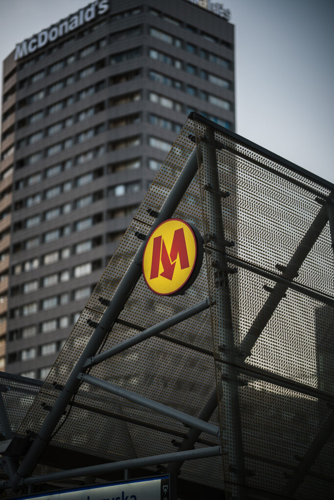 Warsaw Metro "M" sign on a perforated entrance canopy, with a soft-focus tower block rising behind it