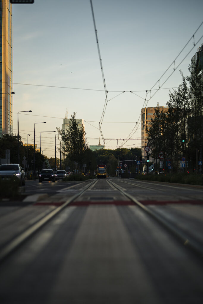 Low view along Warsaw tram tracks at sunset, a yellow tram approaching under overhead wires, foreground rails blurred by the Sigma 85mm F1.2