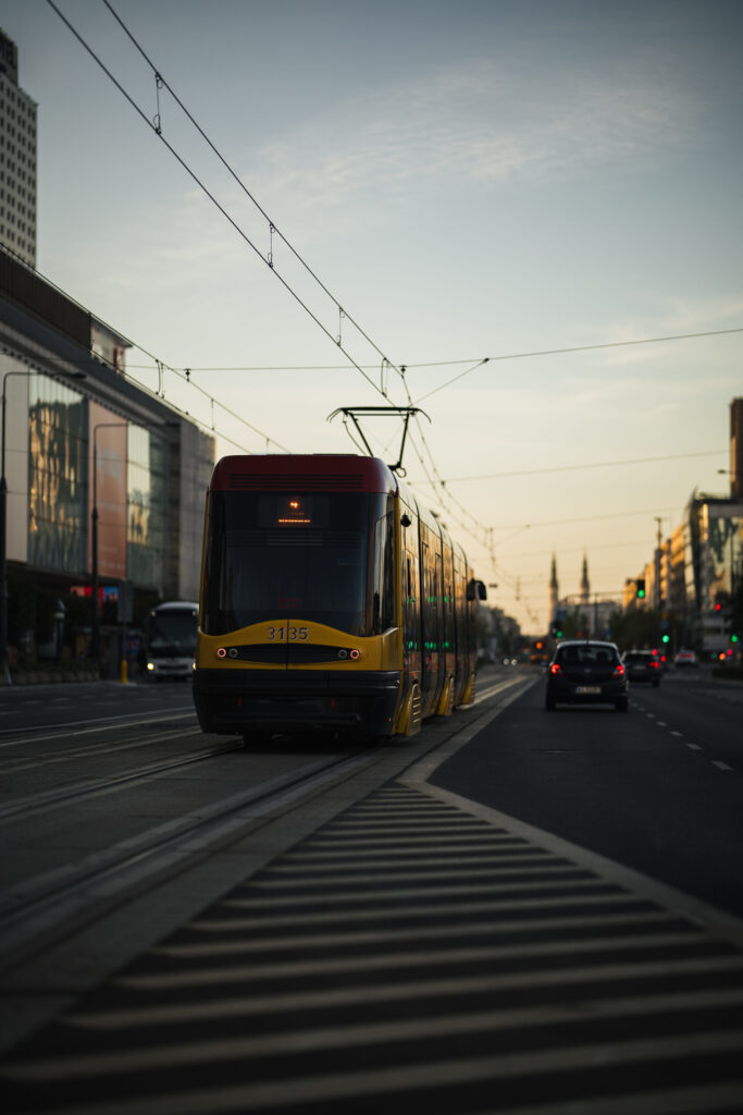 Yellow and red Warsaw tram heading down a wide avenue at dusk, cars alongside and church spires in the distance
